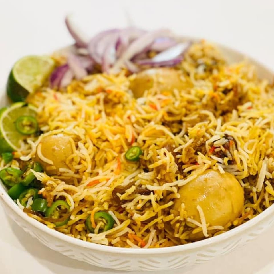 A bowl of Bangladeshi biryani with rice, potatoes, sliced onions, lime, and green chilies.