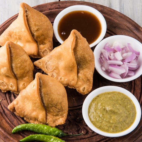 Golden-brown samosas with green chilies, tamarind chutney, mint chutney, and chopped red onions.