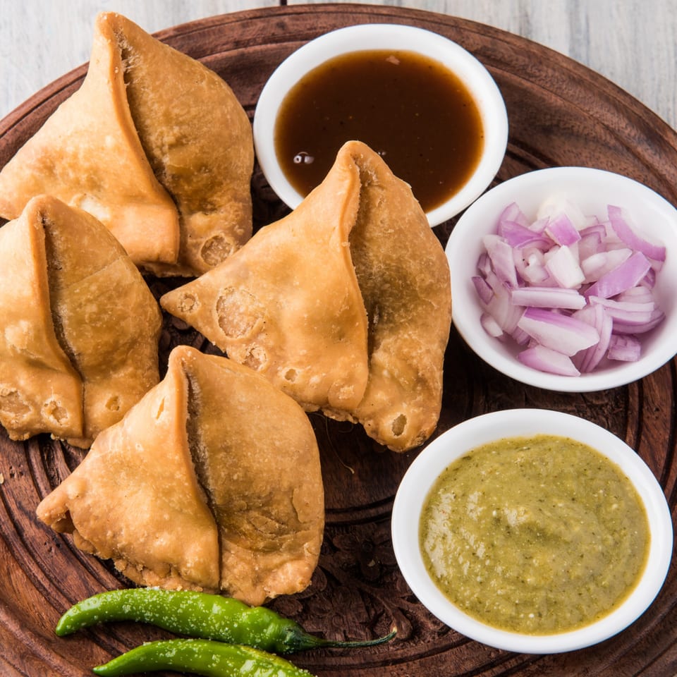 Golden-brown samosas with green chilies, tamarind chutney, mint chutney, and chopped red onions.