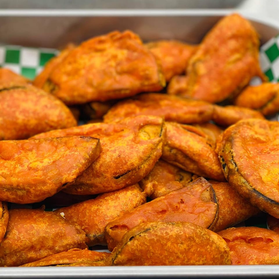 Close-up of many golden-orange Bangladeshi vegetable fritters, possibly begun bhaja, in a white tray with green checkered paper.