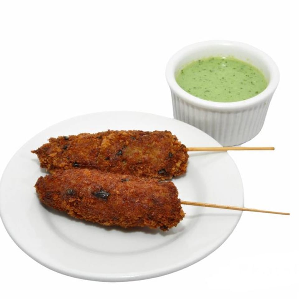 Two Bangladeshi fried seekh kebabs on skewers with a side of green dipping sauce.