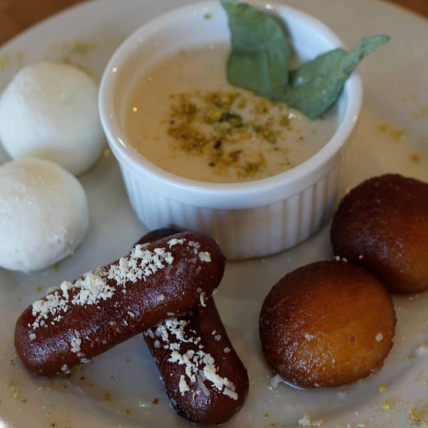 A plate of Bangladeshi desserts, including white rasgulla, gulab jamun, and a creamy pudding with pistachios.