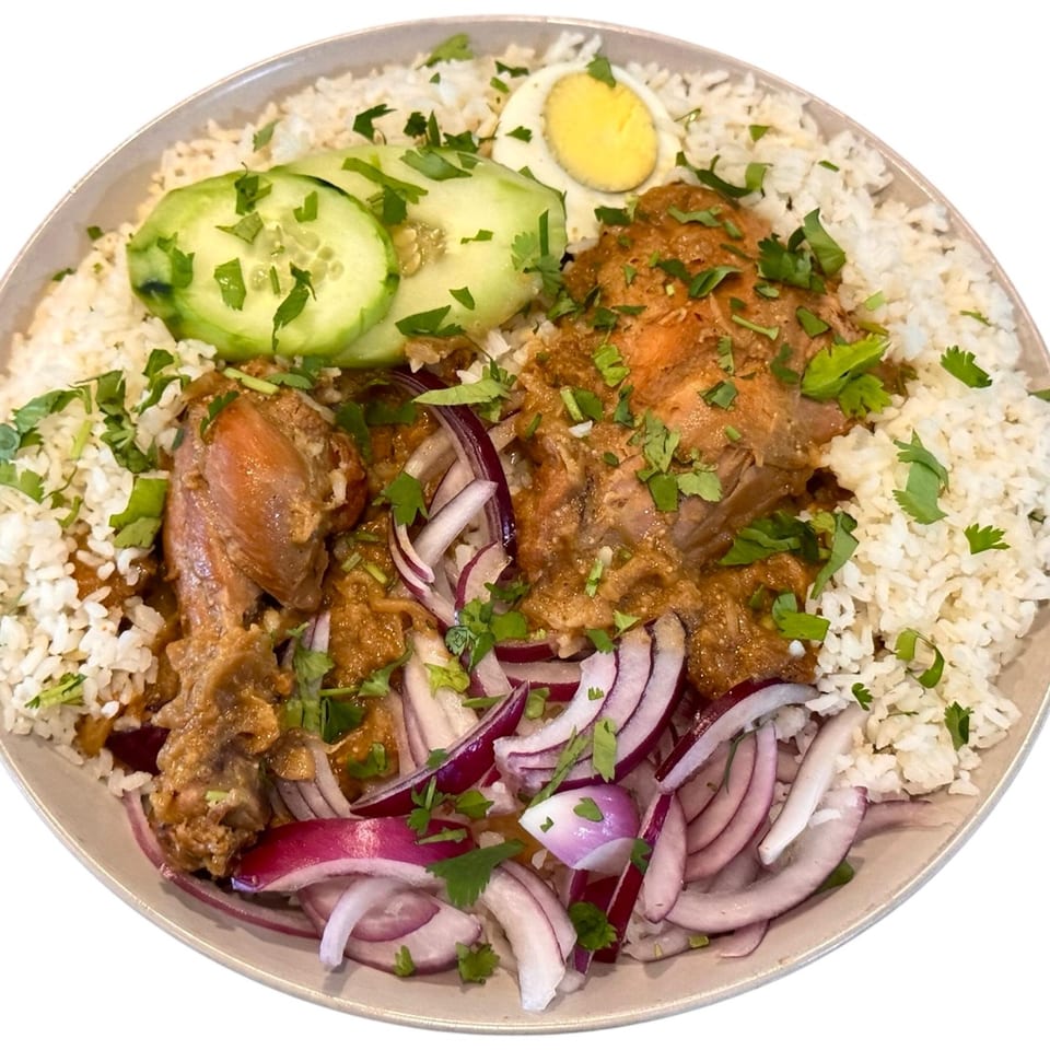 A Bangladeshi dish with chicken curry, white rice, sliced cucumbers, red onions, and a boiled egg.