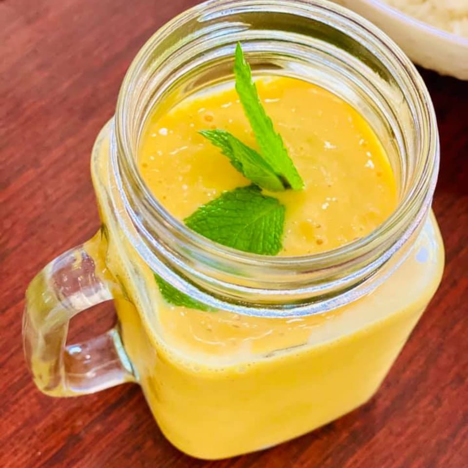 A glass mug of yellow mango lassi, garnished with fresh mint leaves, on a wooden table.