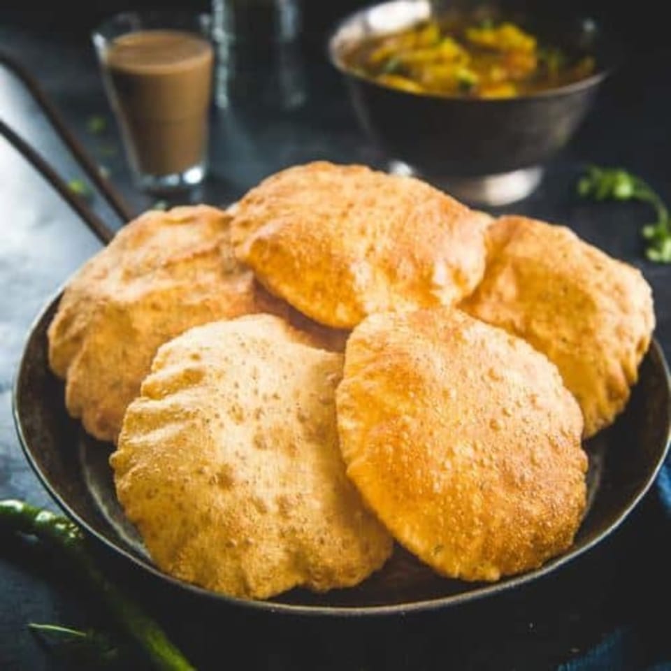 Golden-brown Bangladeshi puri bread in a dark pan with a glass of chai and curry in the background.
