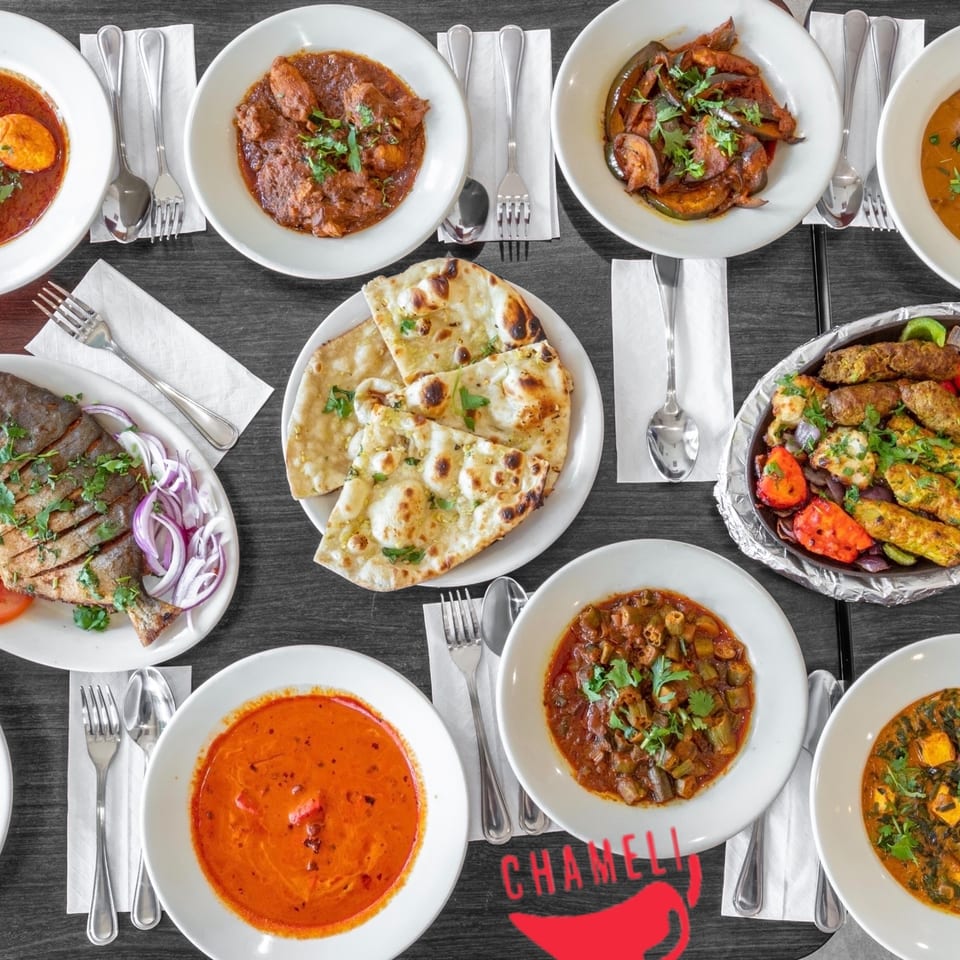 Overhead view of various Bangladeshi dishes, including curries, naan, grilled fish, and kebabs, on a dark table.